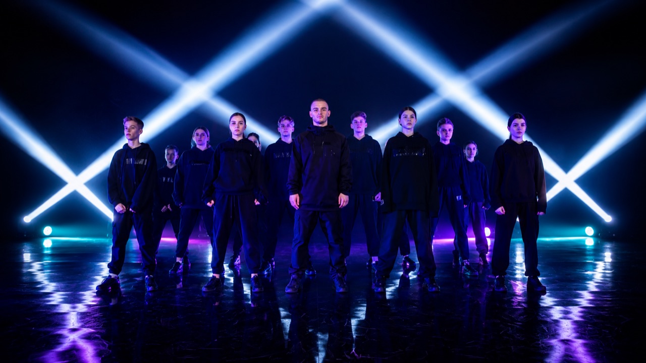 Dance video still — a youth dance crew posing together in a bright studio