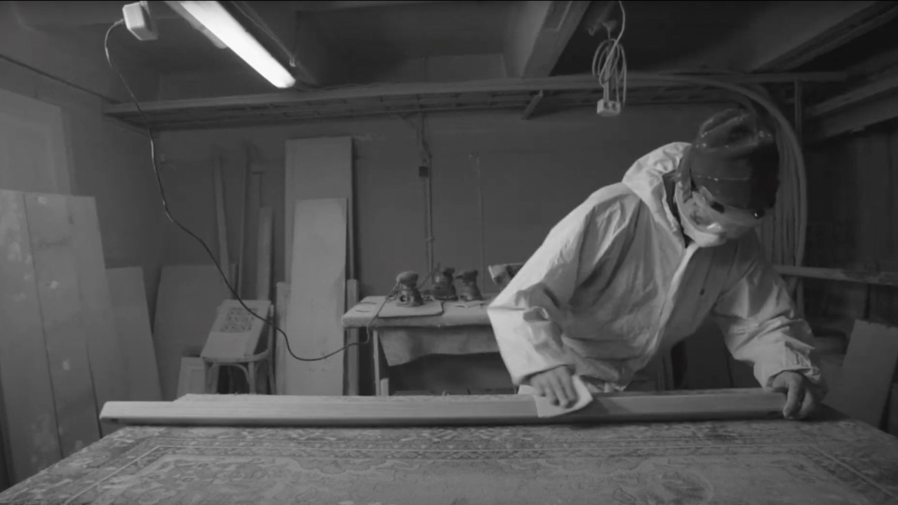 Wood Project documentary still — a woodworker planing lumber in his basement workshop, black and white